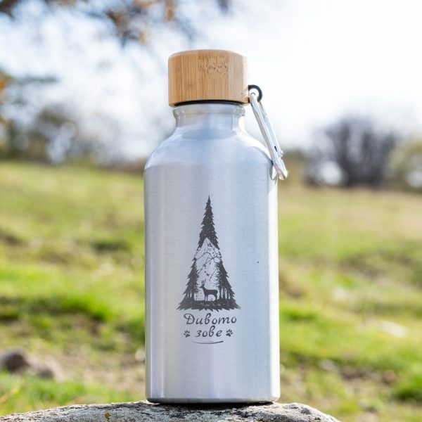 Stainless steel water bottle with bamboo cap and engraved mountain-and-deer design, standing outdoors on a rock.