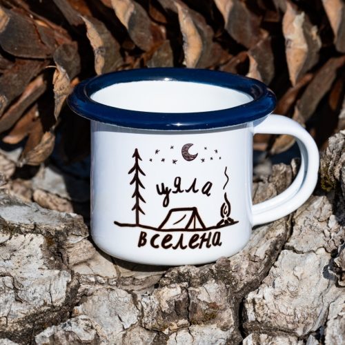 malka-metalna-chasha White enamel mug with blue rim and camping illustration with the text “My Universe”, placed outdoors on bark.