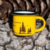 malko-zhylto-kentche Yellow enamel mug with black rim and illustration of pine trees and a tent, with outdoor adventure design, placed on bark.
