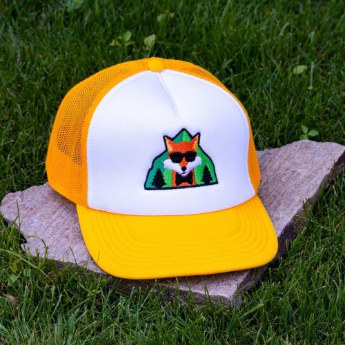 Front view of yellow and white kids mesh cap with embroidered fox wearing sunglasses in front of green mountains, breathable mesh sides and adjustable closure.